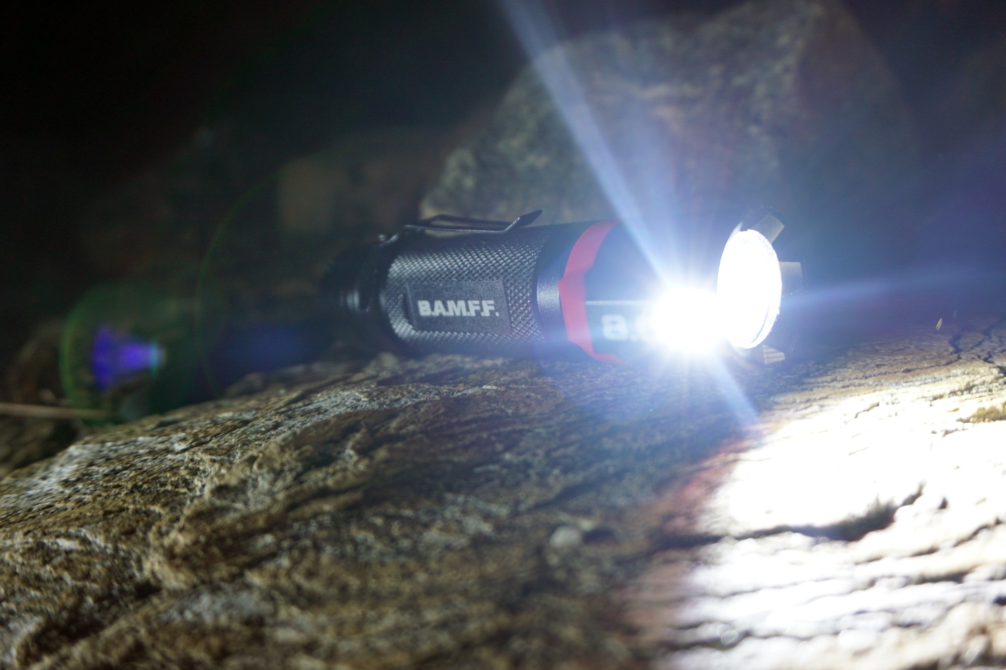 The B.A.M.F.F. Tactical LED flashlight by STKR Concepts sitting on a rock in a night time scene with both flood and spot light on