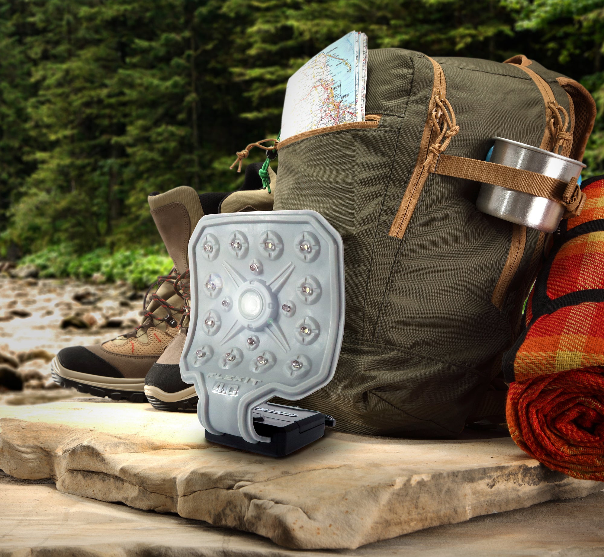 Outdoor image of a hiking backpack spilling out with the essentials like a map, water cup, flashlight, and waterproof boots. All sitting on a rock near a river with a forest full of fur trees in the background.