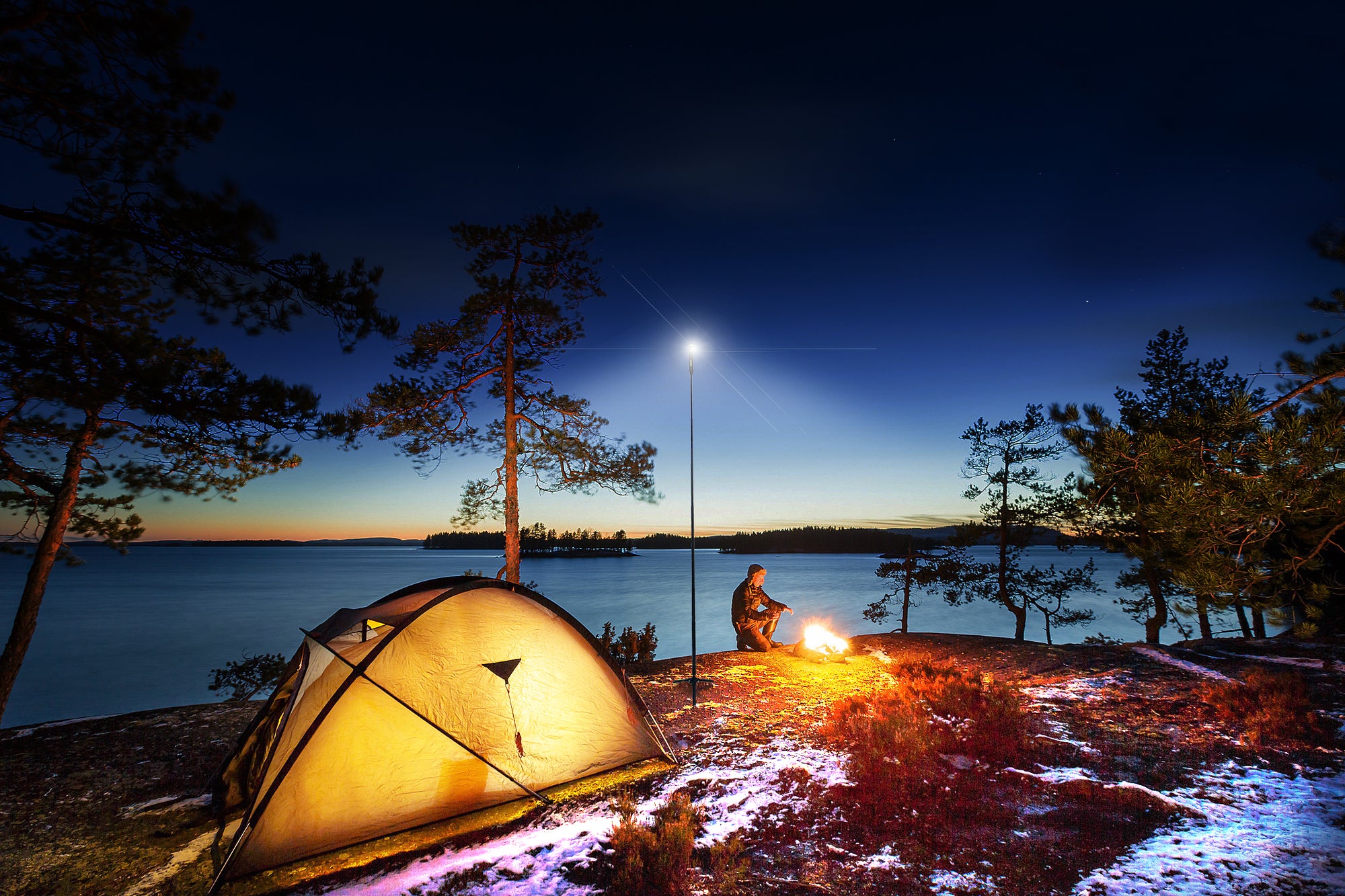Lakeside camping scene featuring a FLi-PRO telescoping area light illuminating a campsite. male figure kneeling by a fire near a tent and some trees