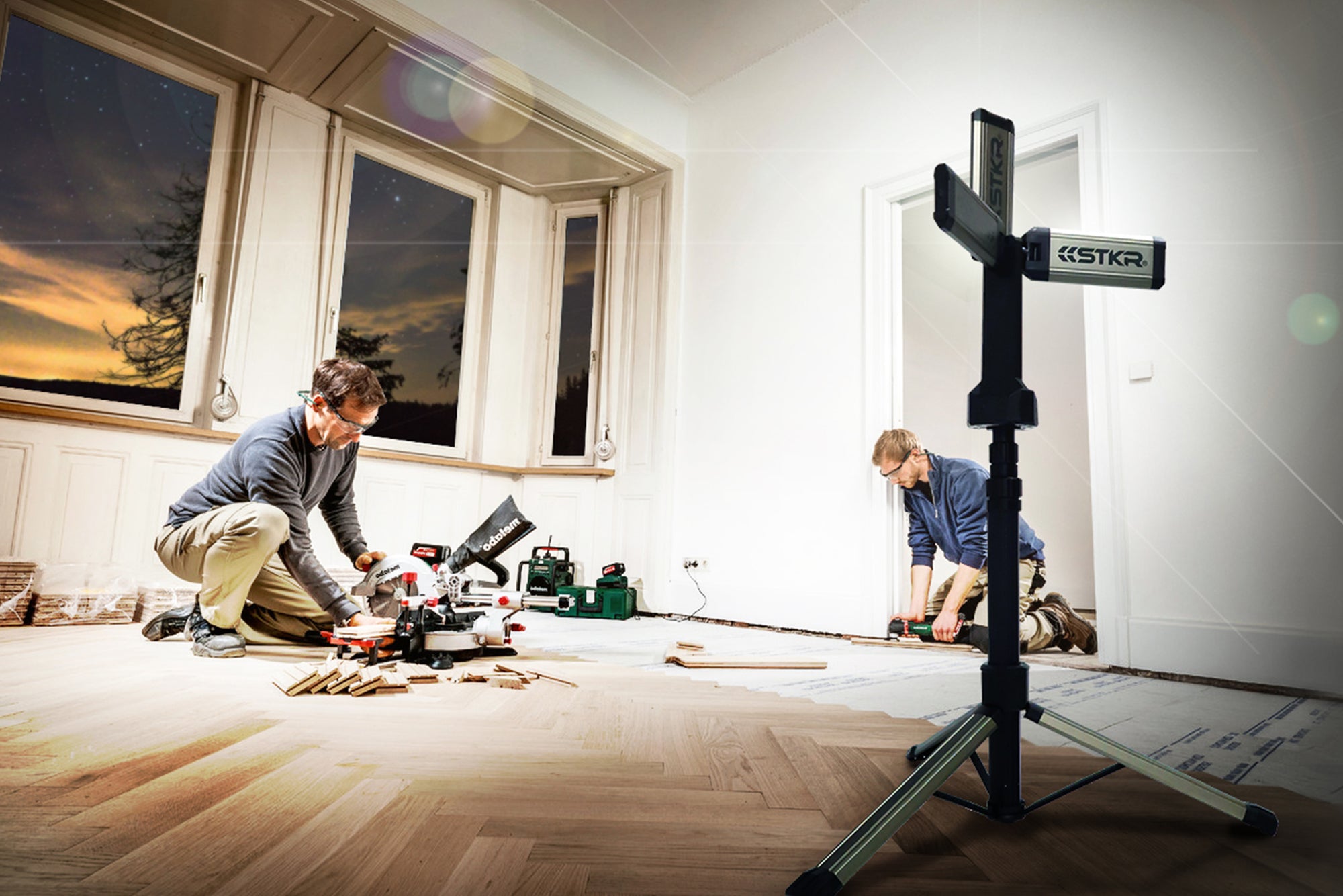 TRi-Mobile area work light on a tripod by STKR Concepts illuminating two male workers in the process of installing new hardwood flooring in a herringbone pattern. DIY home project after dark.