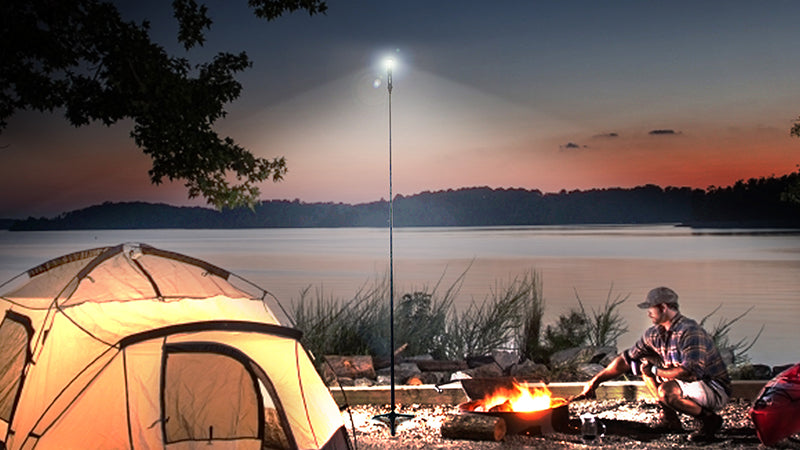 camping scene featuring a male cooking over a fire next to his tent. Nighttime near a lake. He's illuminated by a FLi-PRO telescoping light by STKR Concepts